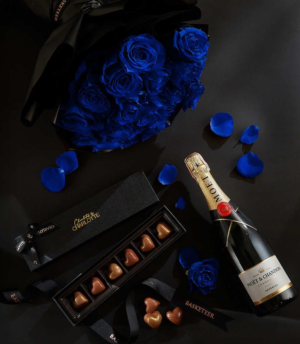 Moët & Chandon champagne bottle with a black box of heart-shaped chocolates, deep blue roses, and rose petals on a black background, with a Basketeer ribbon.