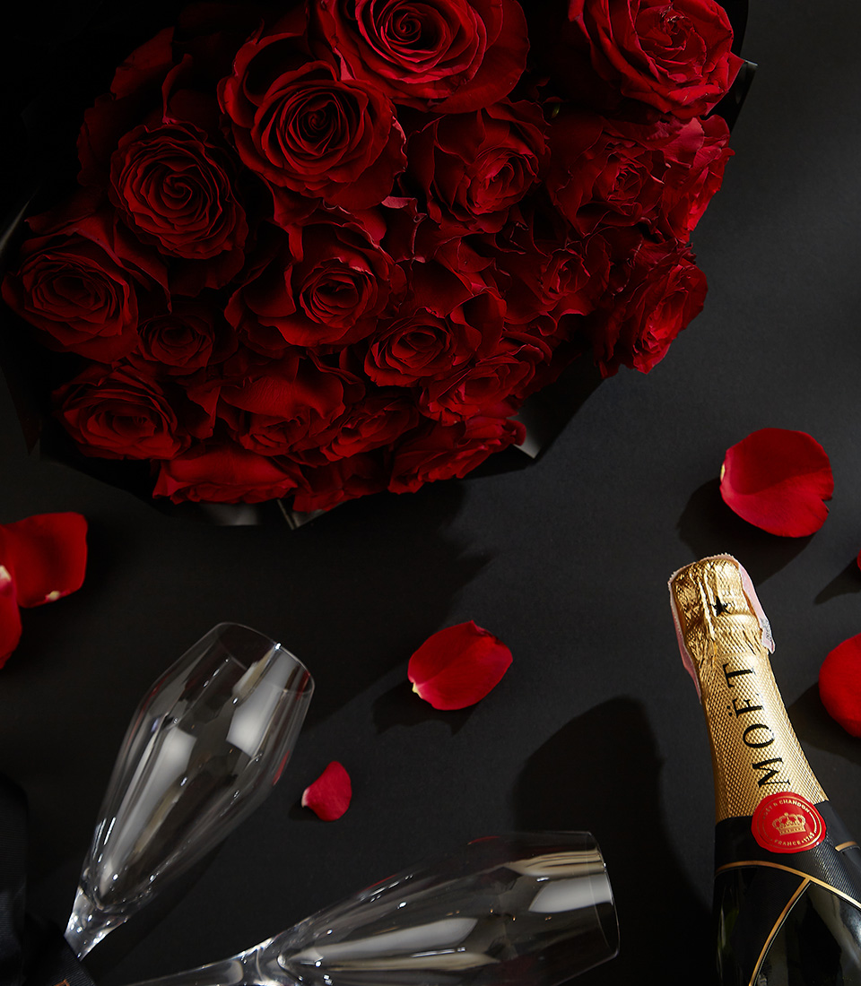 Moët & Chandon champagne bottle with two crystal flutes, surrounded by a bouquet of deep red roses and scattered rose petals on a black background with a Basketeer ribbon.