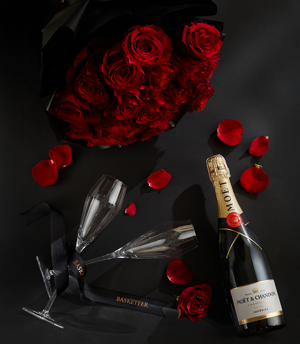 Moët & Chandon champagne bottle with two crystal flutes, surrounded by a bouquet of deep red roses and scattered rose petals on a black background with a Basketeer ribbon.