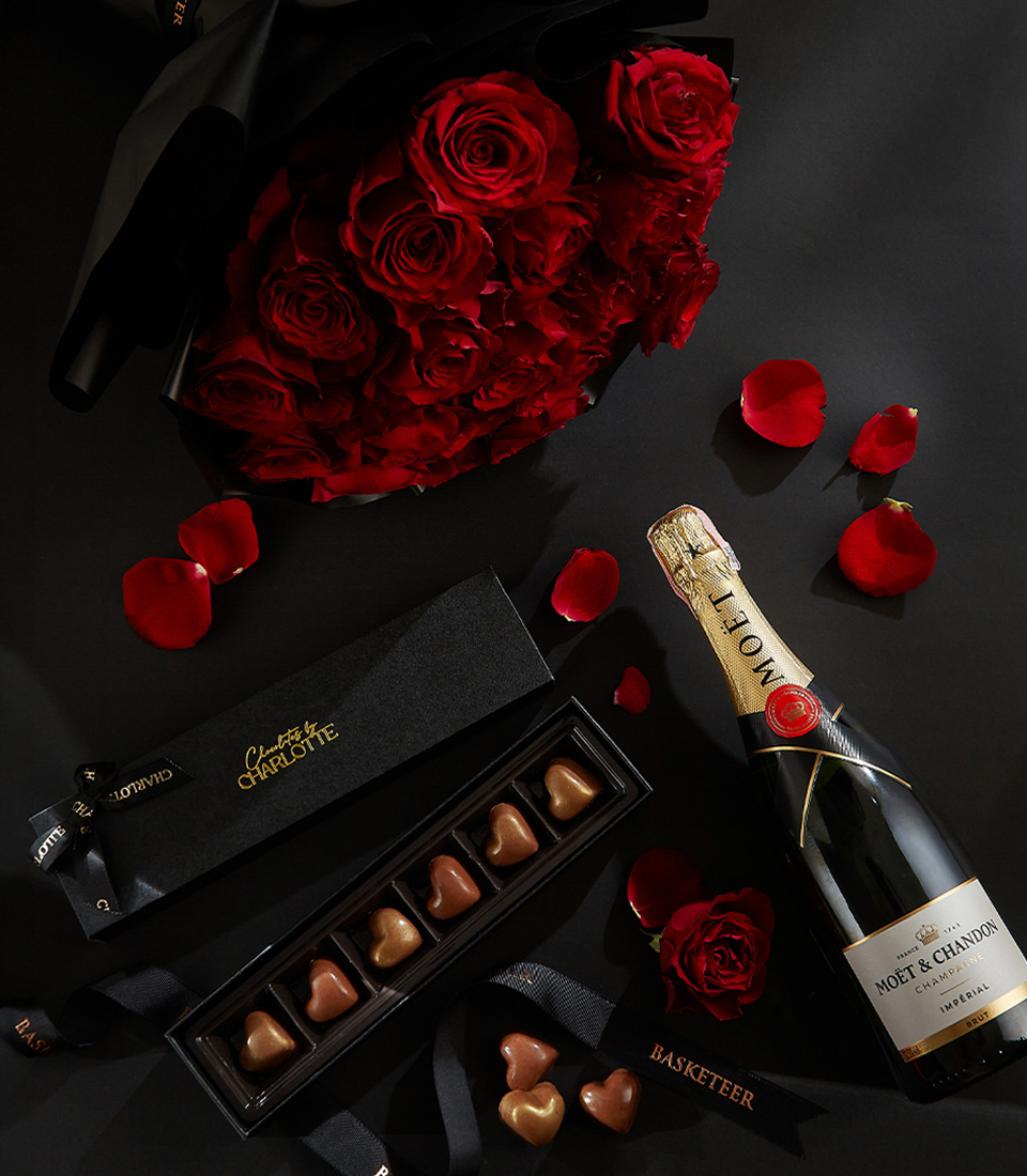 Moët & Chandon champagne bottle, heart-shaped chocolates in a black box, and a bouquet of deep red roses, surrounded by scattered rose petals on a black background with a Basketeer ribbon.