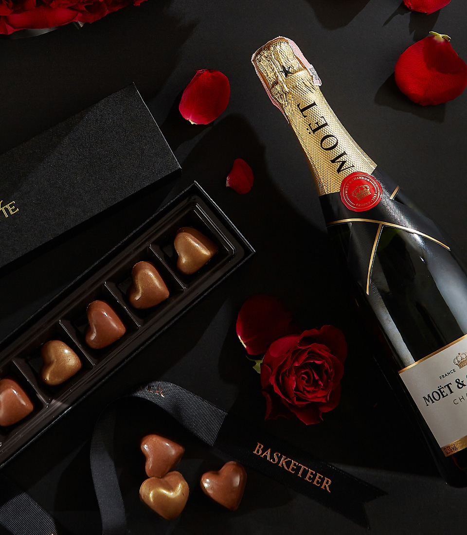 Moët & Chandon champagne bottle, heart-shaped chocolates in a black box, and a bouquet of deep red roses, surrounded by scattered rose petals on a black background with a Basketeer ribbon.