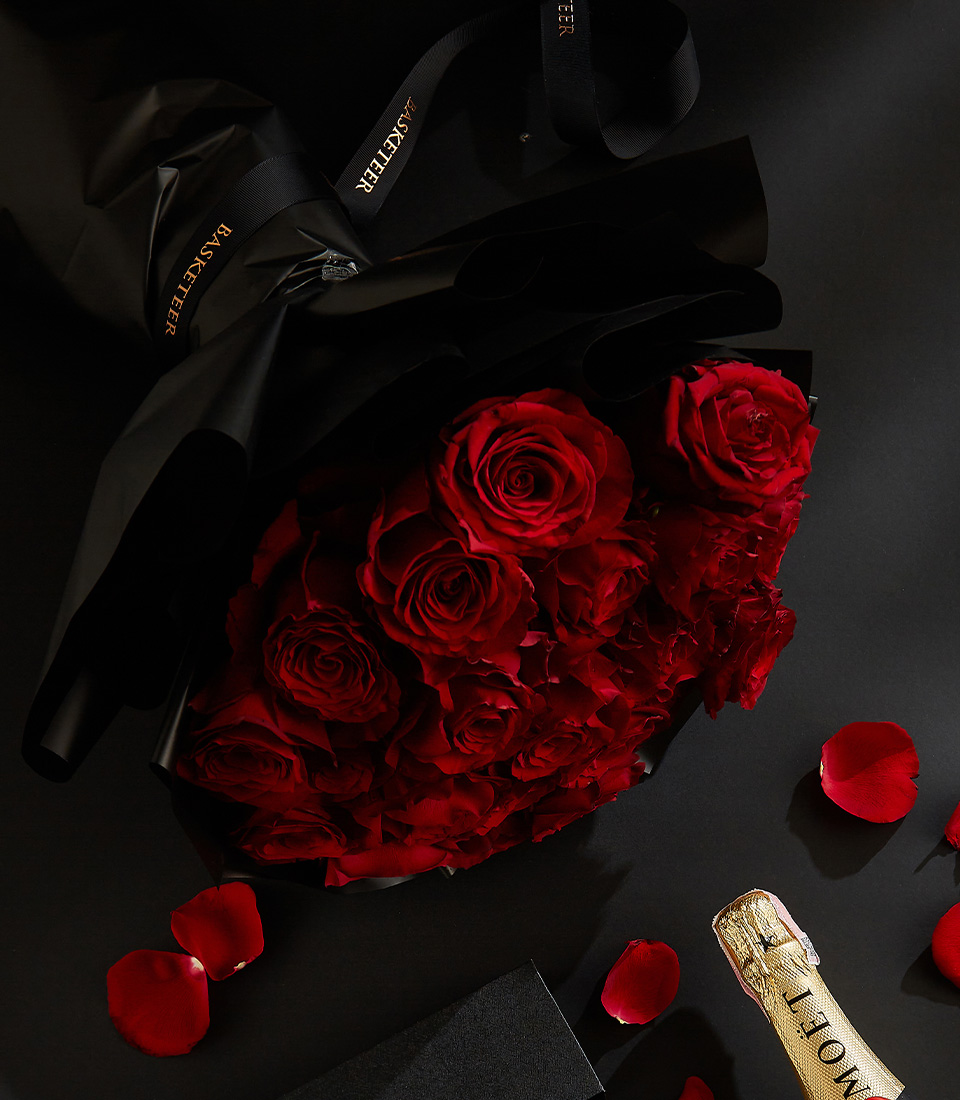 Moët & Chandon champagne bottle, heart-shaped chocolates in a black box, and a bouquet of deep red roses, surrounded by scattered rose petals on a black background with a Basketeer ribbon.