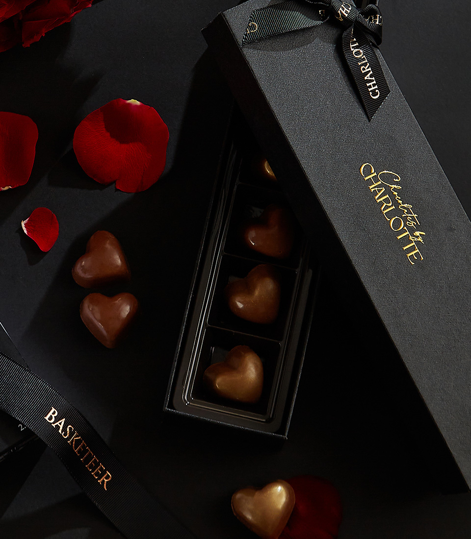 Luxury gift set featuring a bouquet of deep red roses, a designer perfume bottle, and a box of heart-shaped chocolates, beautifully arranged on a black background with scattered rose petals.