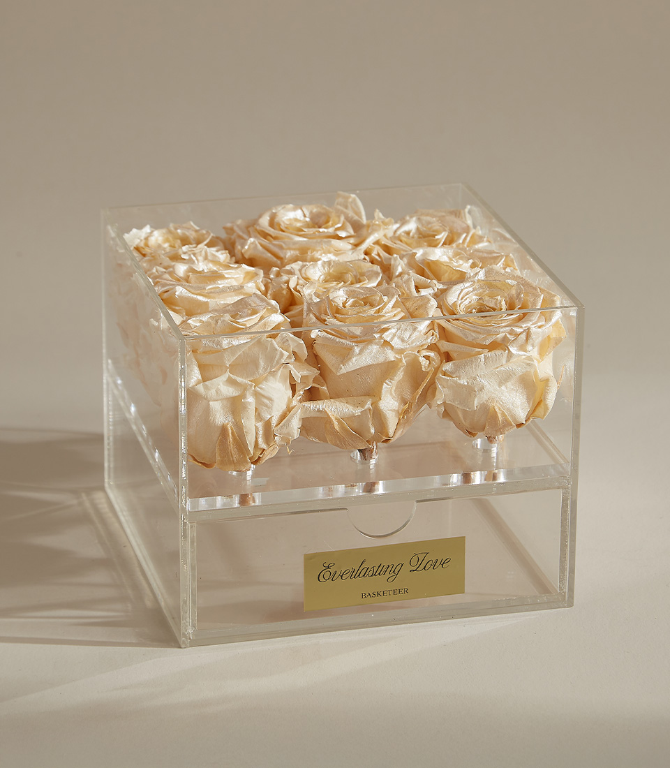 A box of nine champagne gold preserved roses in a clear acrylic display case, accented with a white satin ribbon and an elegant gold plaque reading 