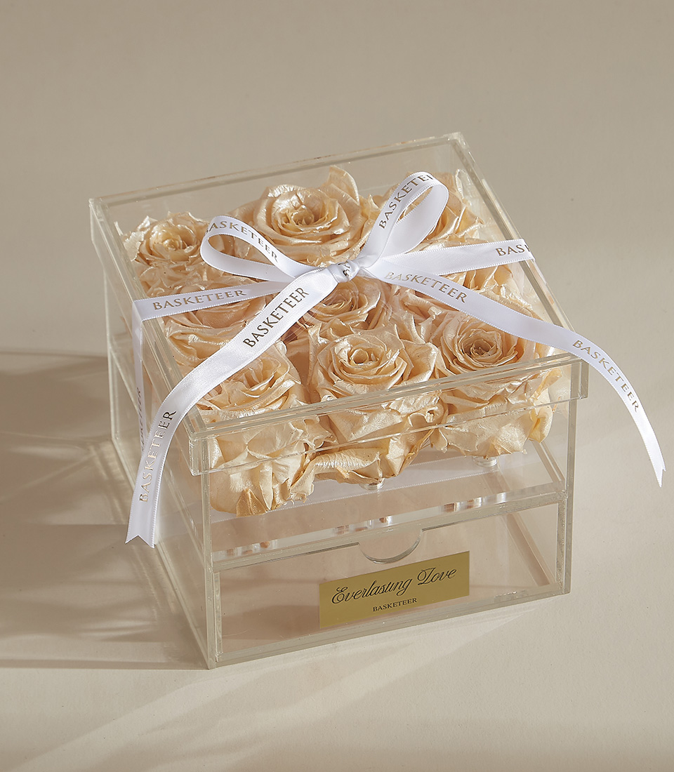 A box of nine champagne gold preserved roses in a clear acrylic display case, accented with a white satin ribbon and an elegant gold plaque reading 