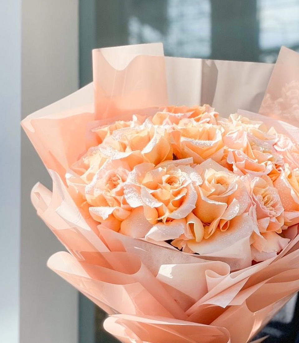 Close-up of a stunning bouquet of grand peach roses with a soft shimmer, elegantly wrapped in peach-toned paper, held by a person wearing black.