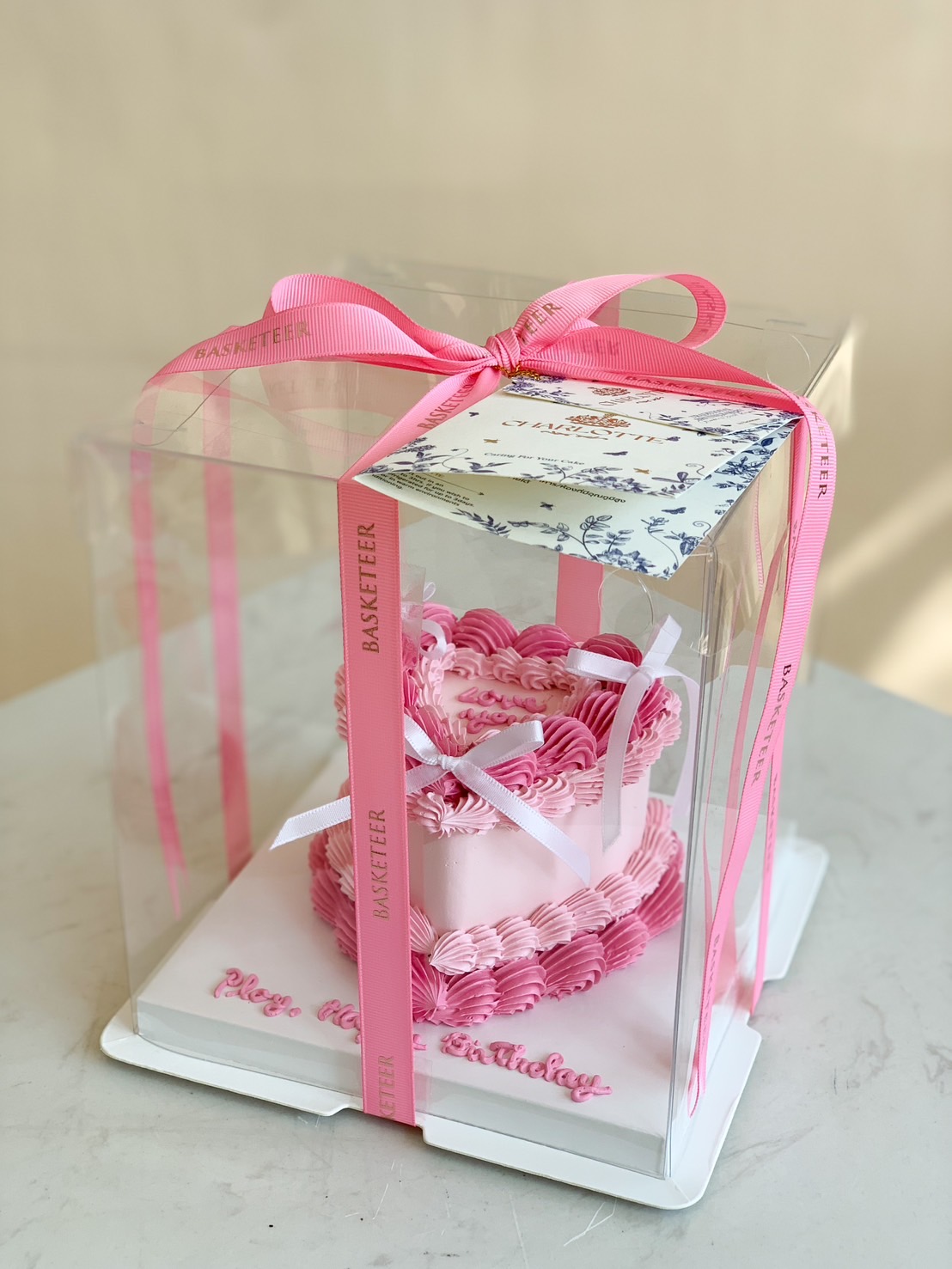 Pink ombre heart-shaped birthday cake with delicate ruffle buttercream frosting, elegantly packed in a clear gift box with a pink satin ribbon.