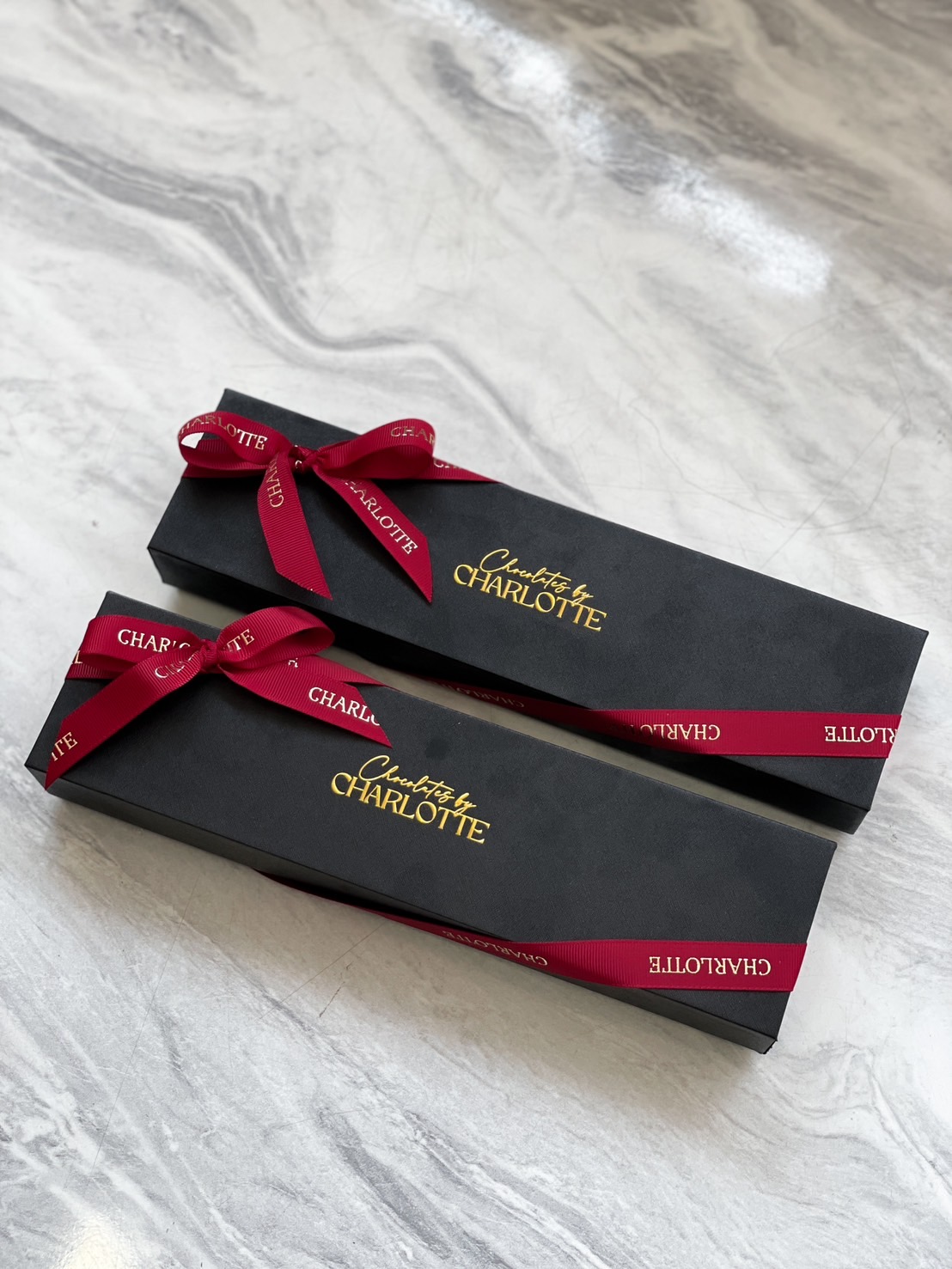 Luxurious black gift boxes adorned with gold “Chocolates by Charlotte” and signature red ribbons on elegant marble in Bangkok.
