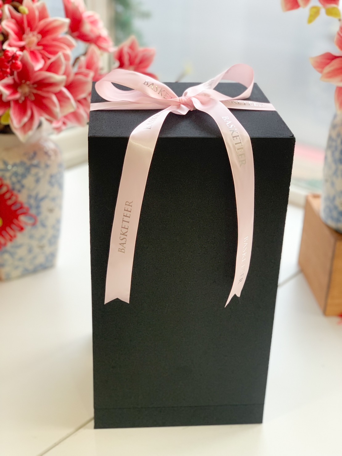A luxury black gift box with a satin Basketeer ribbon, placed in front of elegant preserved rose arrangements in soft pastel tones.