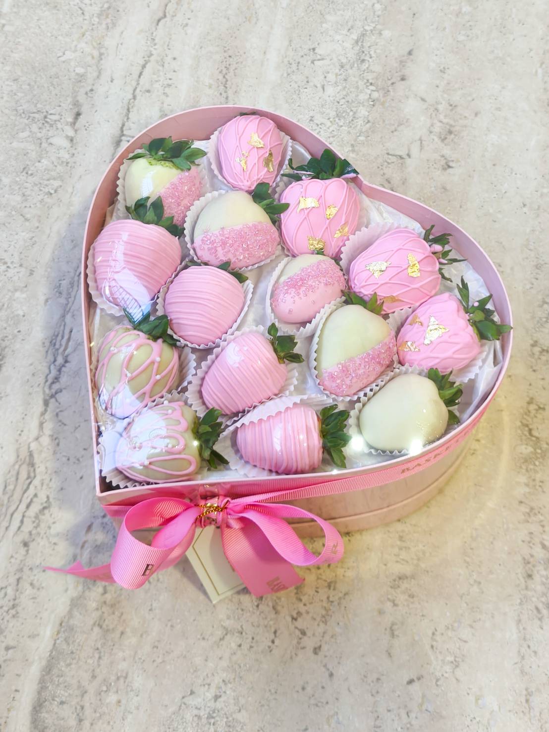 Heart-shaped gift box filled with pink and white chocolate-covered strawberries decorated with gold leaf and sprinkles.