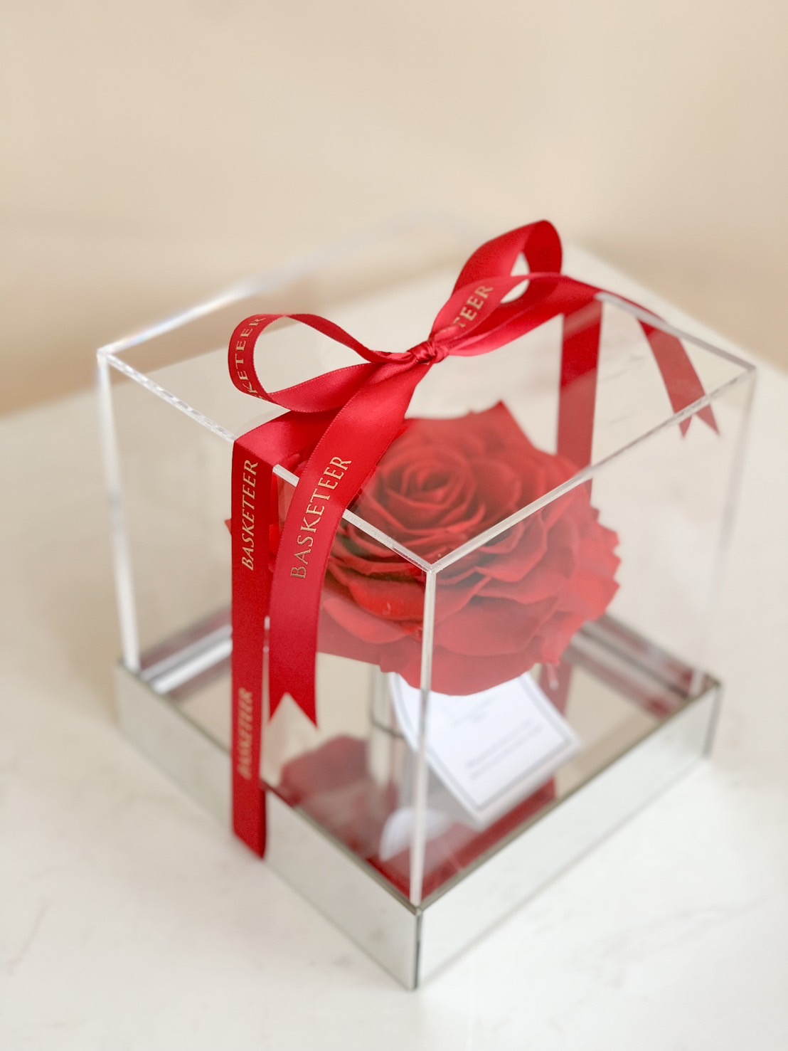 A single red preserved rose displayed in a clear acrylic box with a reflective base, tied with a red Basketeer ribbon.