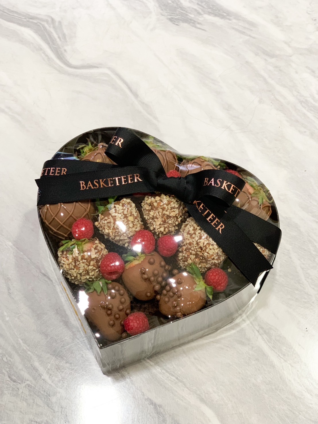 A heart-shaped box filled with chocolate-dipped strawberries, topped with hazelnut, chocolate pearls, and drizzles, paired with fresh raspberries, and tied with a black Basketeer ribbon.