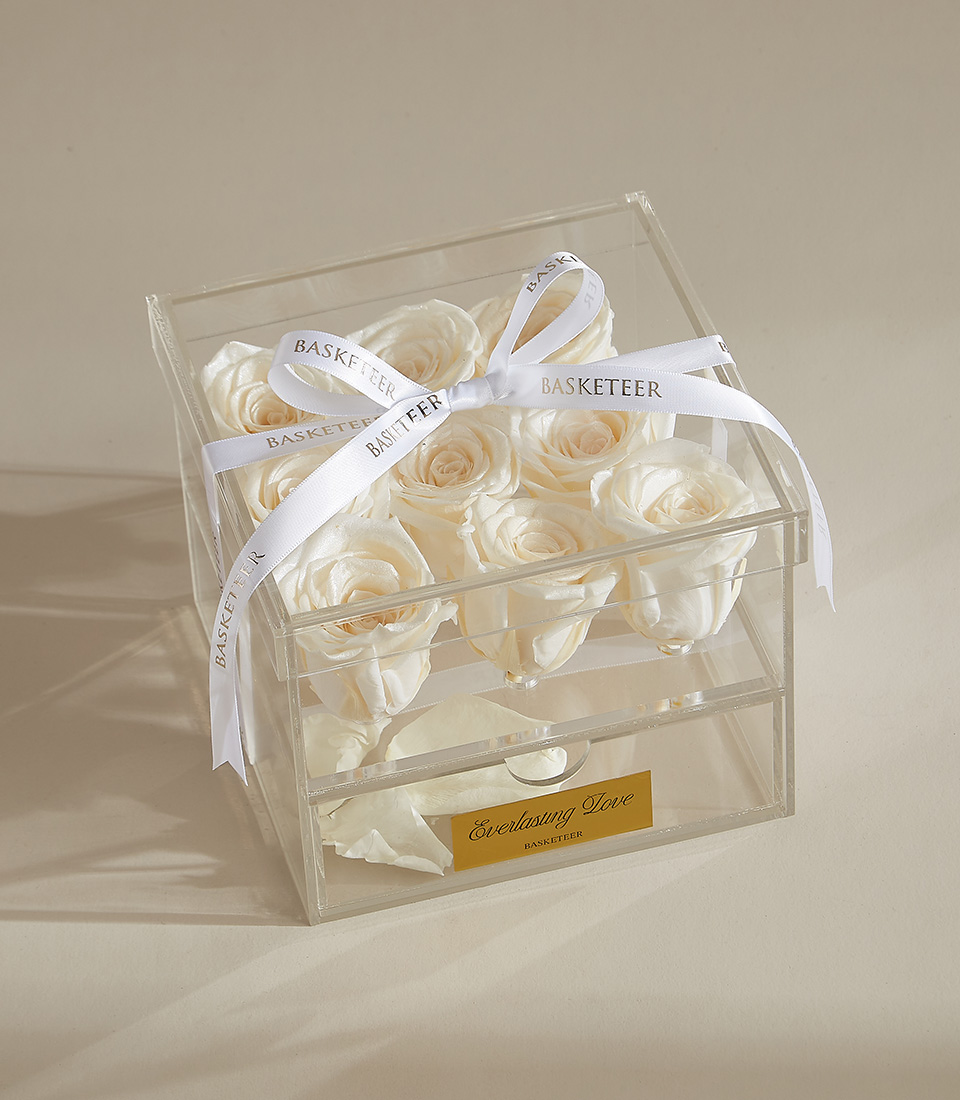 A luxurious preserved white rose arrangement featuring nine elegant roses in a clear acrylic box with a keepsake drawer, tied with a white ribbon.