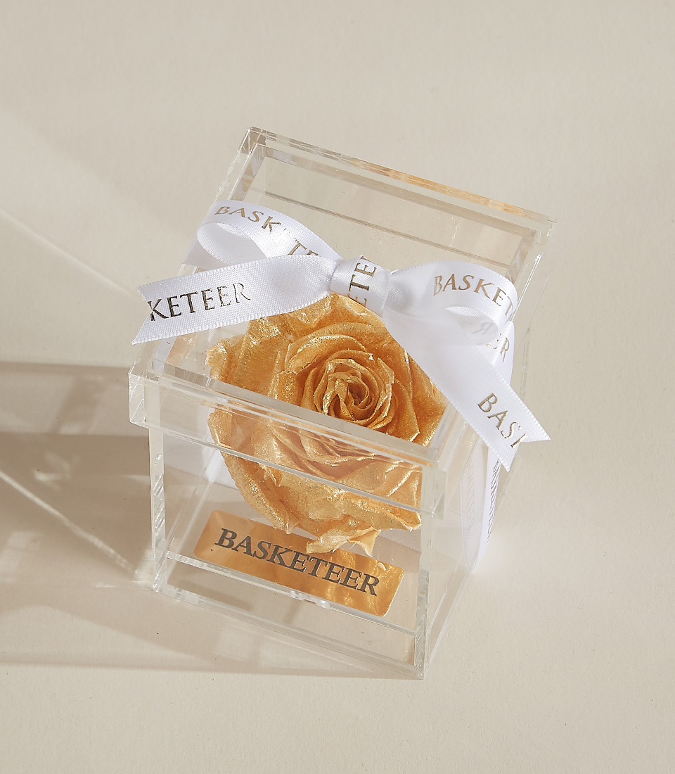 A golden preserved rose beautifully placed in a transparent acrylic box with a gold-engraved Basketeer nameplate and an elegant white ribbon.