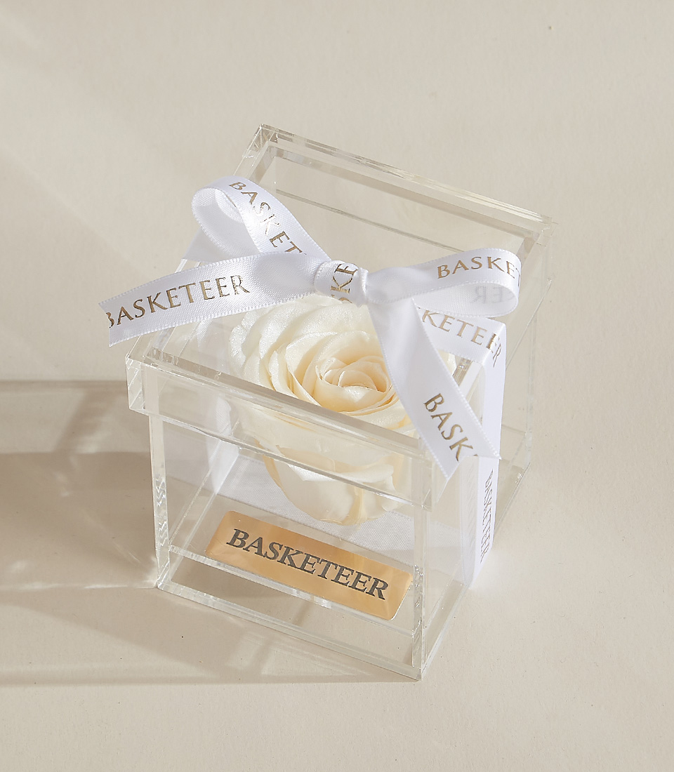 A white preserved rose elegantly placed in a transparent acrylic box with a gold-engraved Basketeer nameplate and a delicate white ribbon.