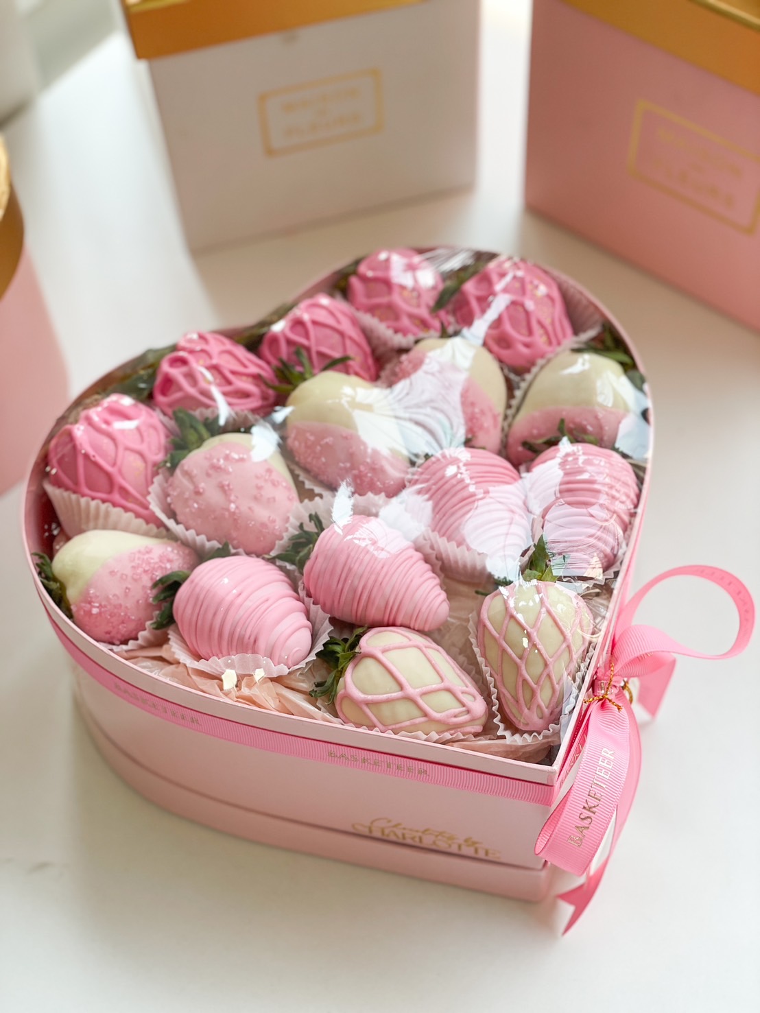 A luxurious heart-shaped gift box filled with pink and white chocolate-covered strawberries, elegantly decorated with drizzle and sprinkles, wrapped with a pink ribbon.