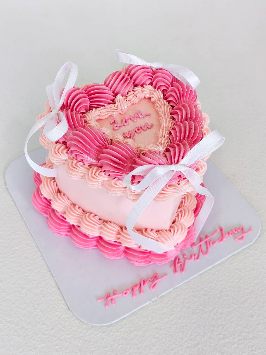 Heart-shaped pink birthday cake with piped cream design, white ribbon bows, and “Love You” message on top.