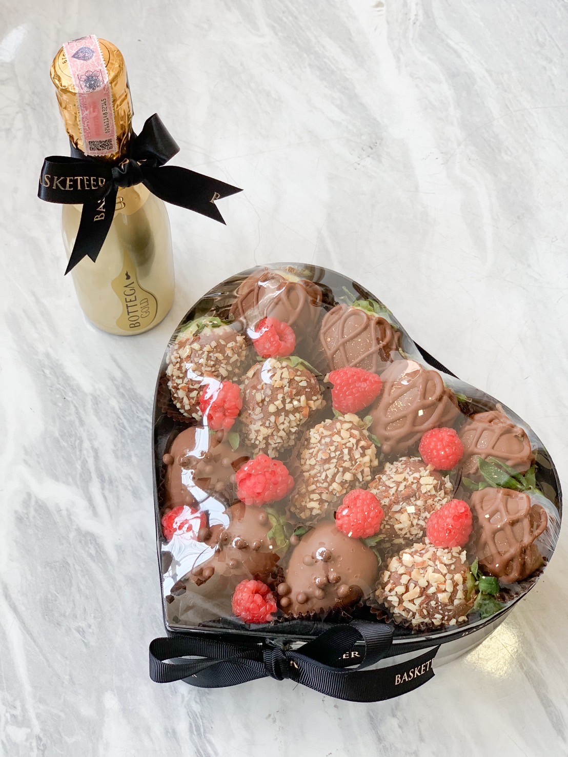 A heart-shaped box of dark chocolate strawberries and raspberries, adorned with nuts and a chocolate drizzle, rests on a marble surface. Beside it, a Bottega Gold bottle wrapped in a black bow adds an elegant touch to the indulgent scene.