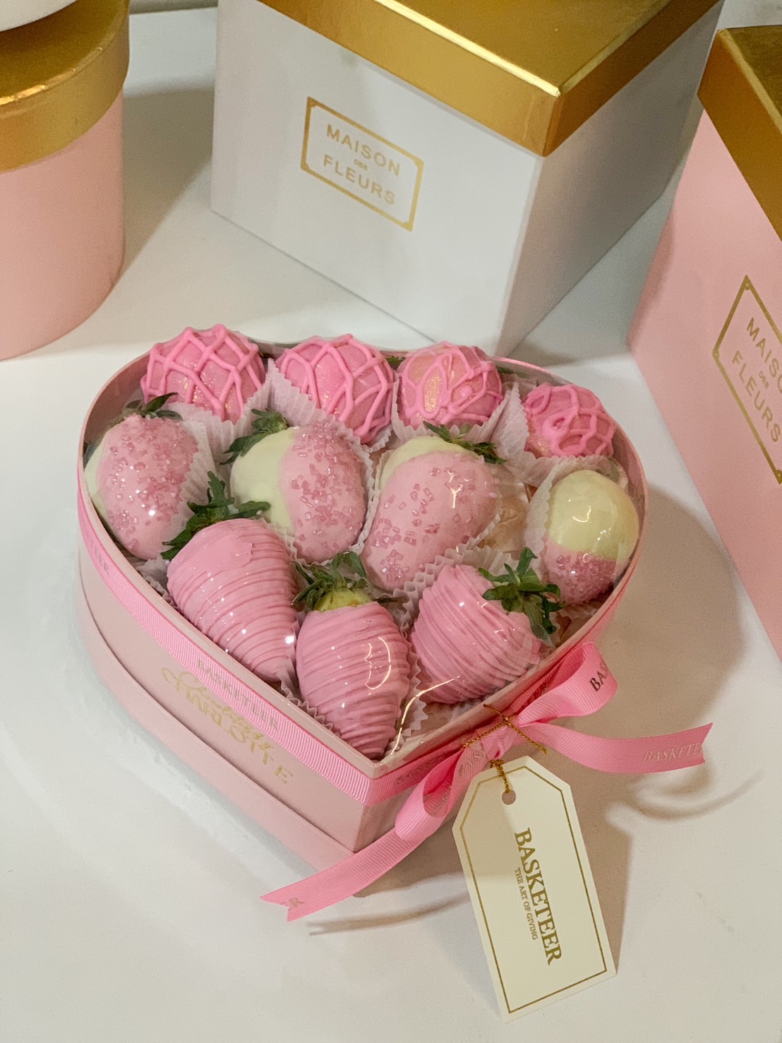 A luxurious heart-shaped gift box filled with pink and white chocolate-covered strawberries, elegantly decorated with drizzle and sprinkles, wrapped with a pink ribbon.