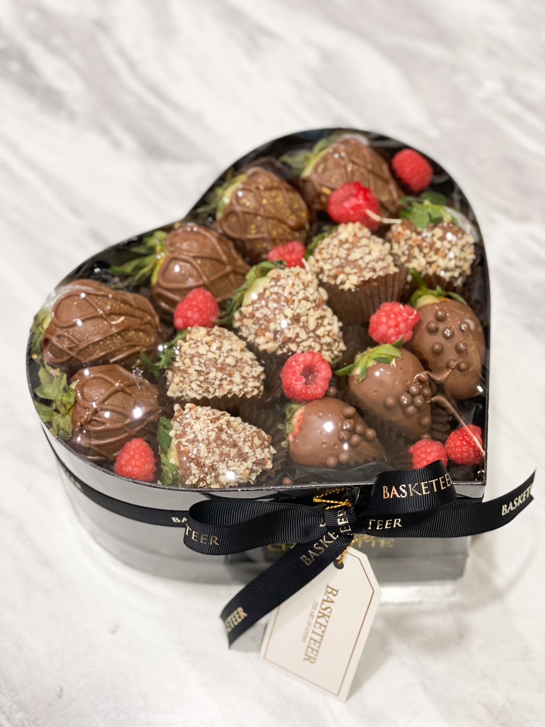 A heart-shaped box filled with chocolate-dipped strawberries, topped with hazelnut, chocolate pearls, and drizzles, paired with fresh raspberries, and tied with a black Basketeer ribbon.