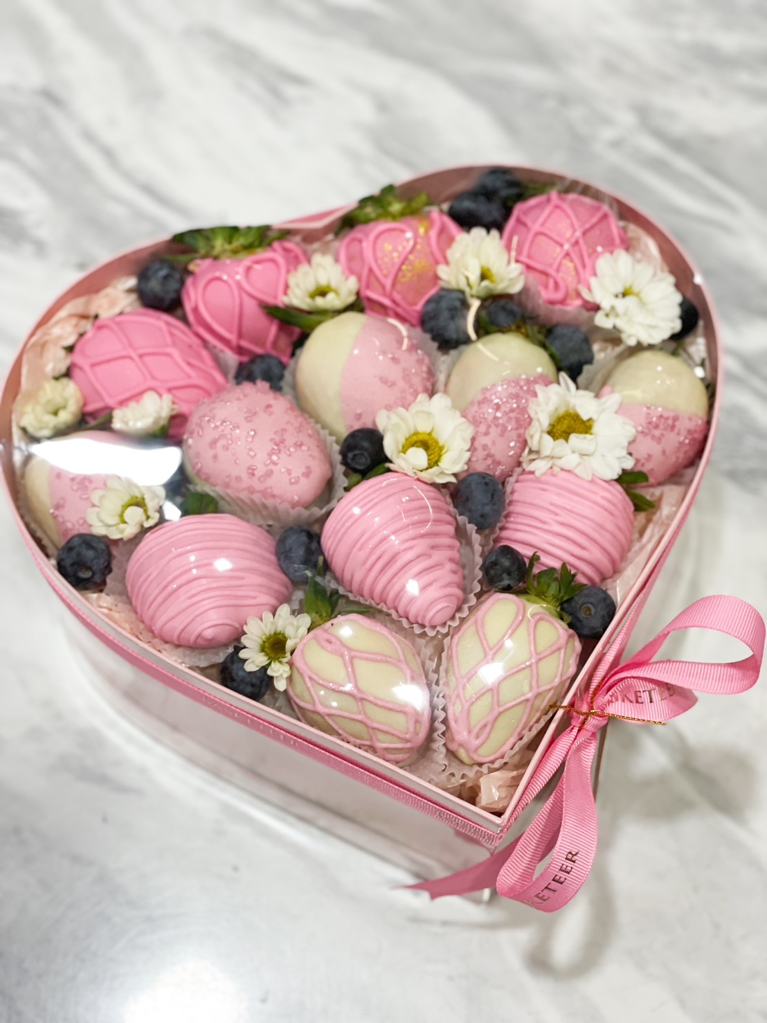 A heart-shaped pink box filled with pink and white chocolate-covered strawberries, blueberries, and small white flowers, creating an elegant and romantic dessert gift.