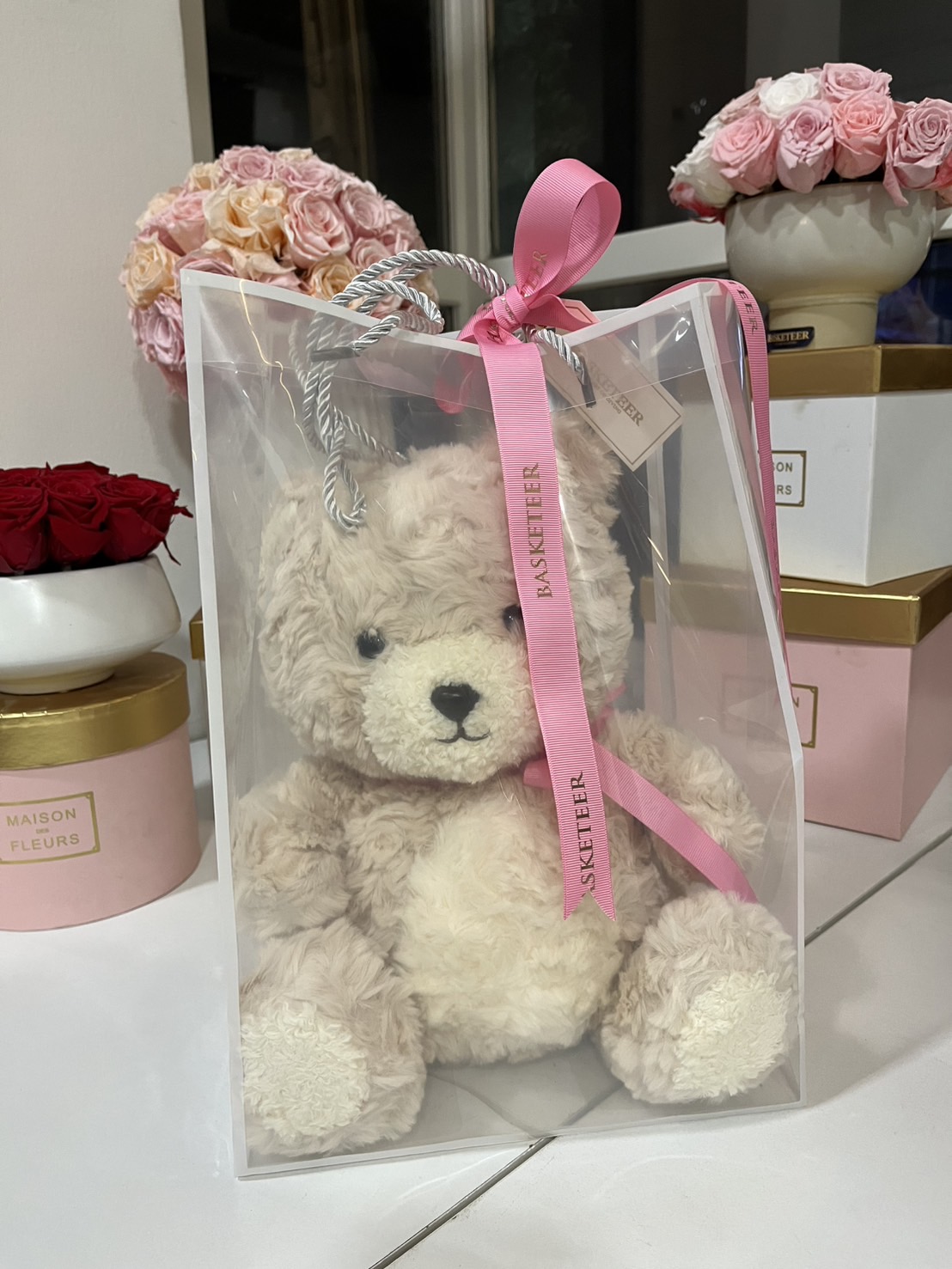 A soft beige teddy bear inside a luxury transparent gift bag, tied with a pink ribbon, placed in an elegant gift setting.