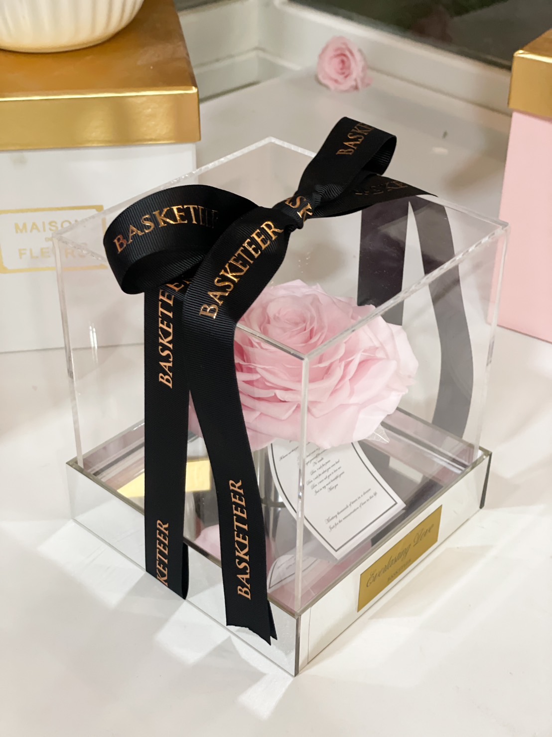 A preserved pink rose elegantly presented in a clear acrylic box with a black satin ribbon imprinted with gold lettering.