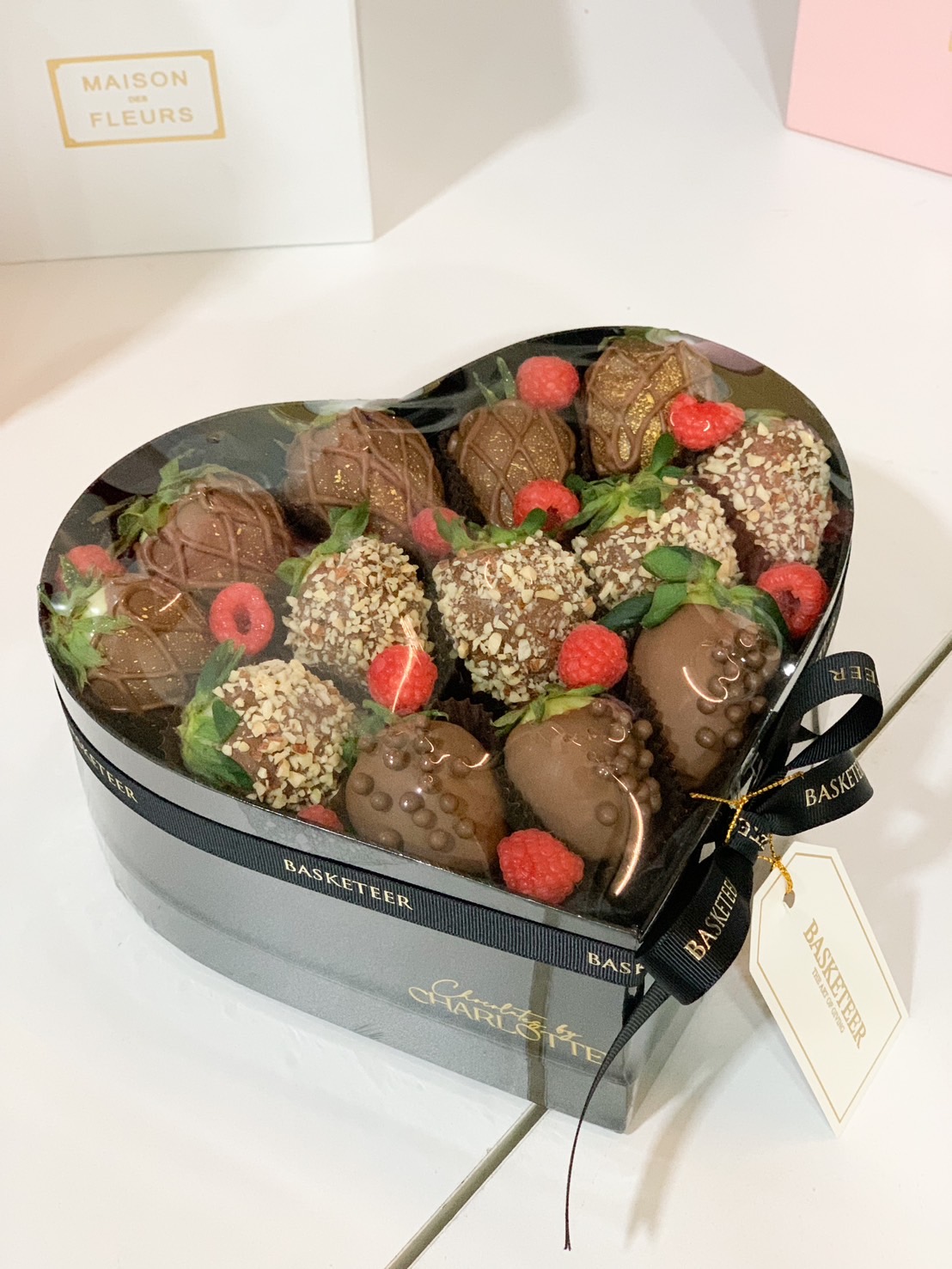 A heart-shaped box filled with chocolate-dipped strawberries, topped with hazelnut, chocolate pearls, and drizzles, paired with fresh raspberries, and tied with a black Basketeer ribbon.