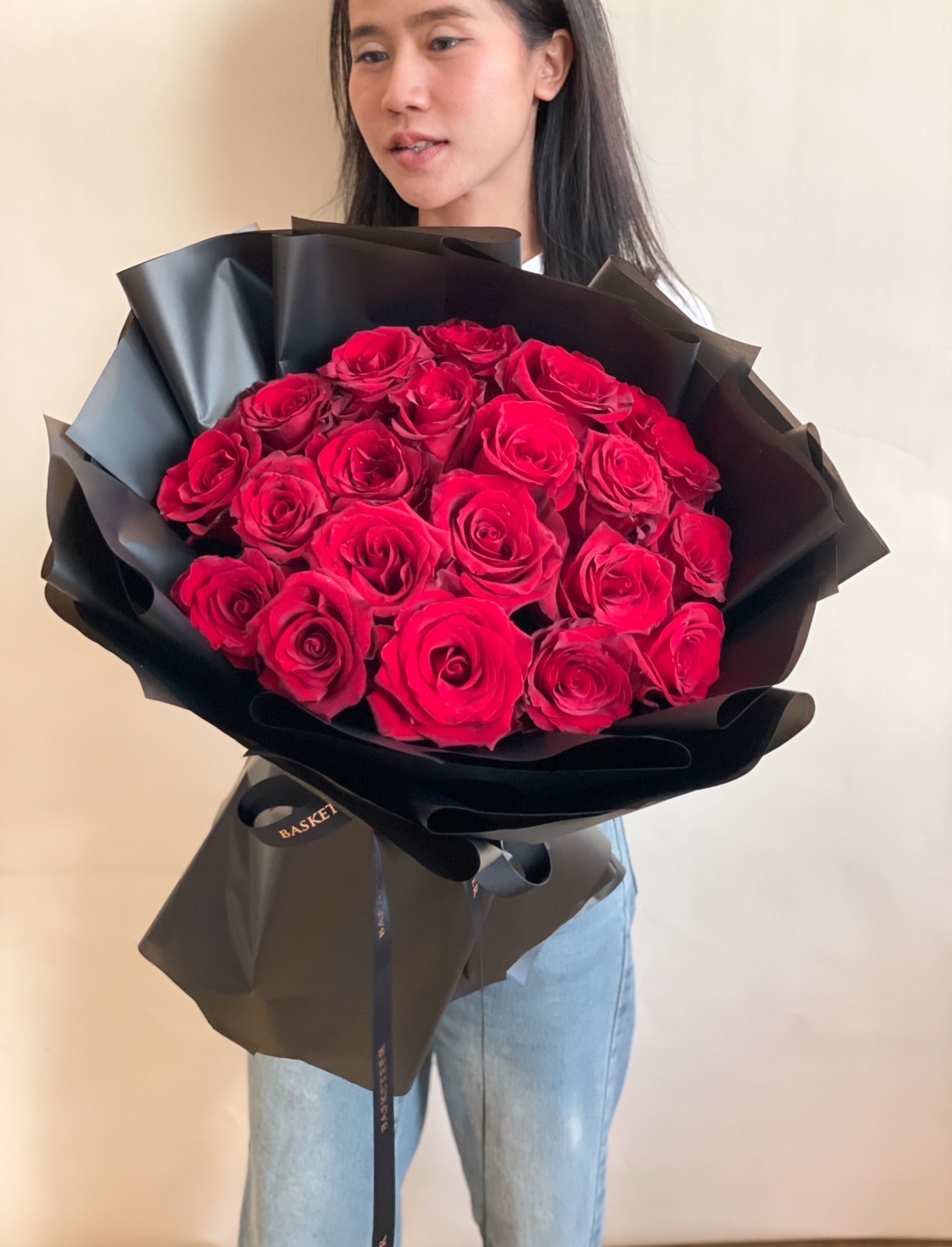 A luxurious bouquet of vibrant red roses wrapped in elegant black paper, held by a woman in a white top and jeans.