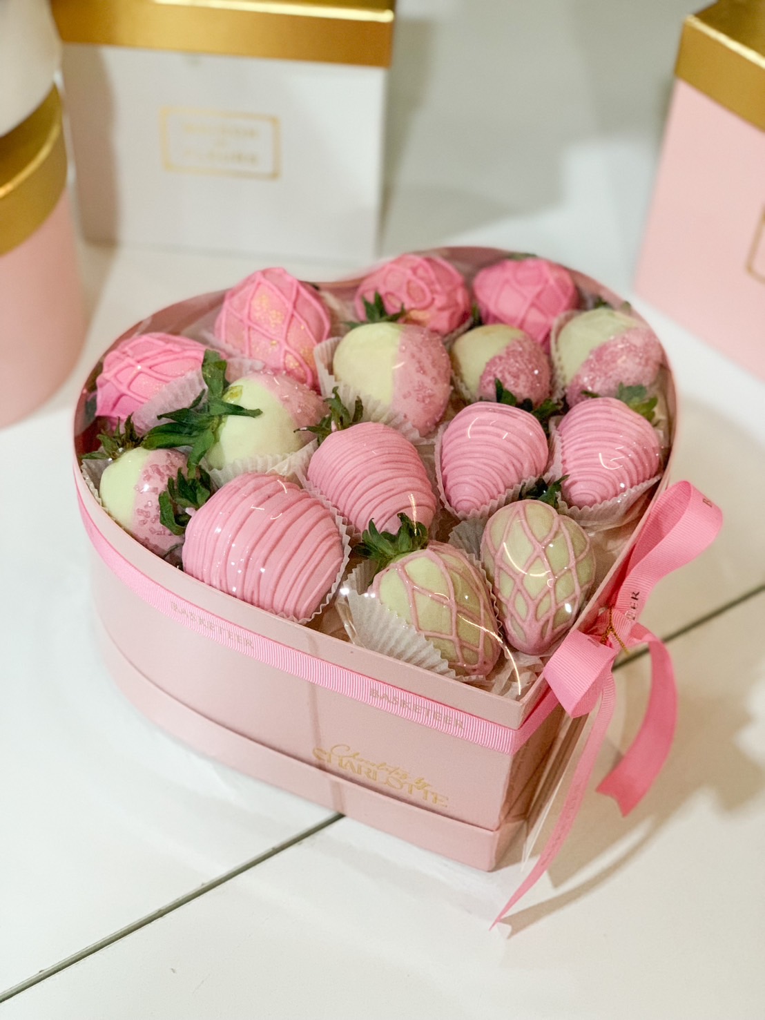 A luxurious heart-shaped gift box filled with pink and white chocolate-covered strawberries, elegantly decorated with drizzle and sprinkles, wrapped with a pink ribbon.