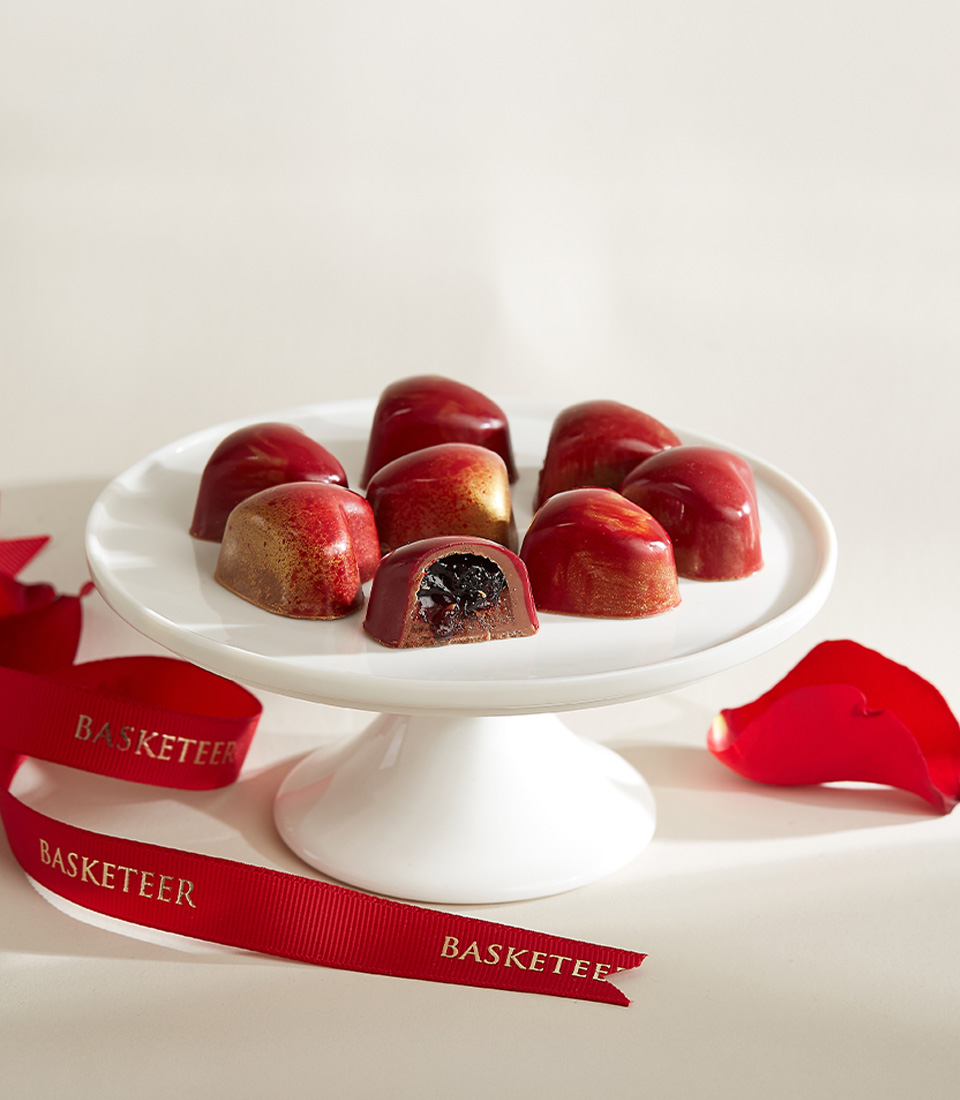 Handcrafted dark cherry chocolates with a glossy red and gold finish, featuring a rich cherry filling, elegantly displayed on a white cake stand.