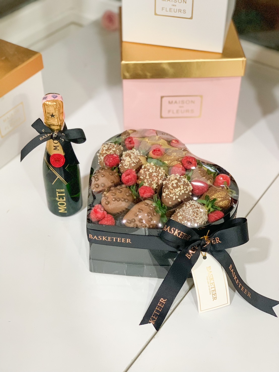 A heart-shaped box of gourmet chocolate-dipped strawberries, adorned with crushed nuts, chocolate drizzle, and fresh raspberries, paired with a Mini Moët champagne bottle, elegantly wrapped with Basketeer branding.