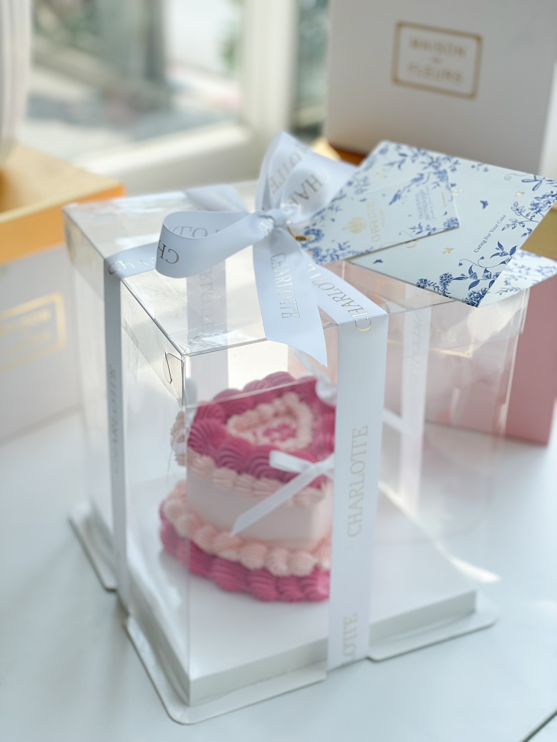 Pink ombre heart-shaped birthday cake with delicate ruffle buttercream frosting, elegantly packed in a clear gift box with a pink satin ribbon.