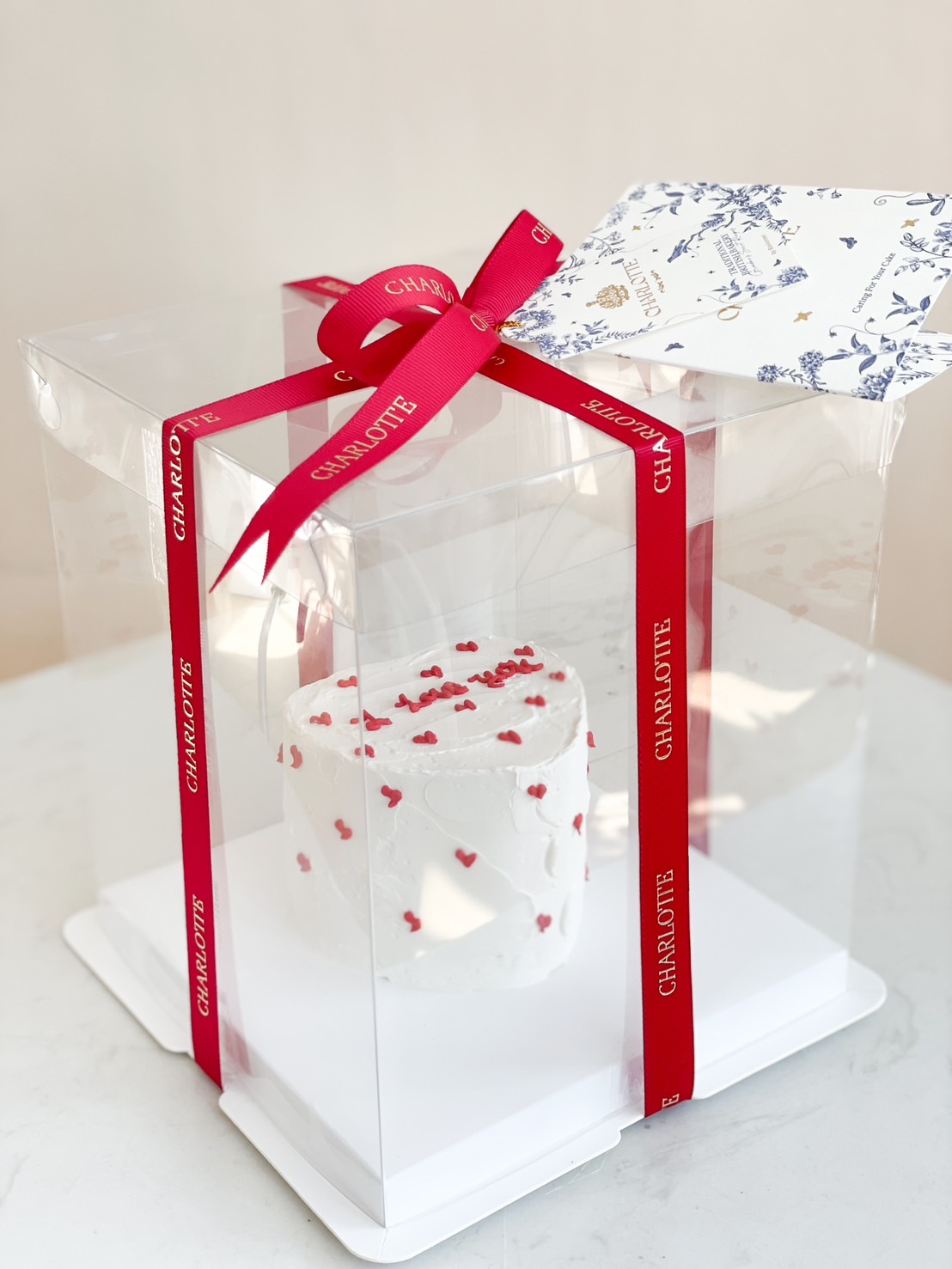 A mini white buttercream cake adorned with red heart confetti, presented in a luxury clear gift box with a red ribbon.