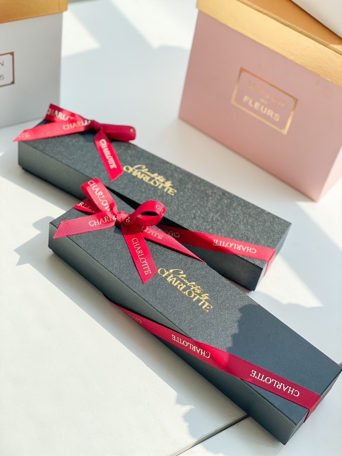 Elegant dark Charlotte Bakery gift boxes with gold lettering and red ribbons beside pastel boxes, bathed in soft Bangkok sunlight.