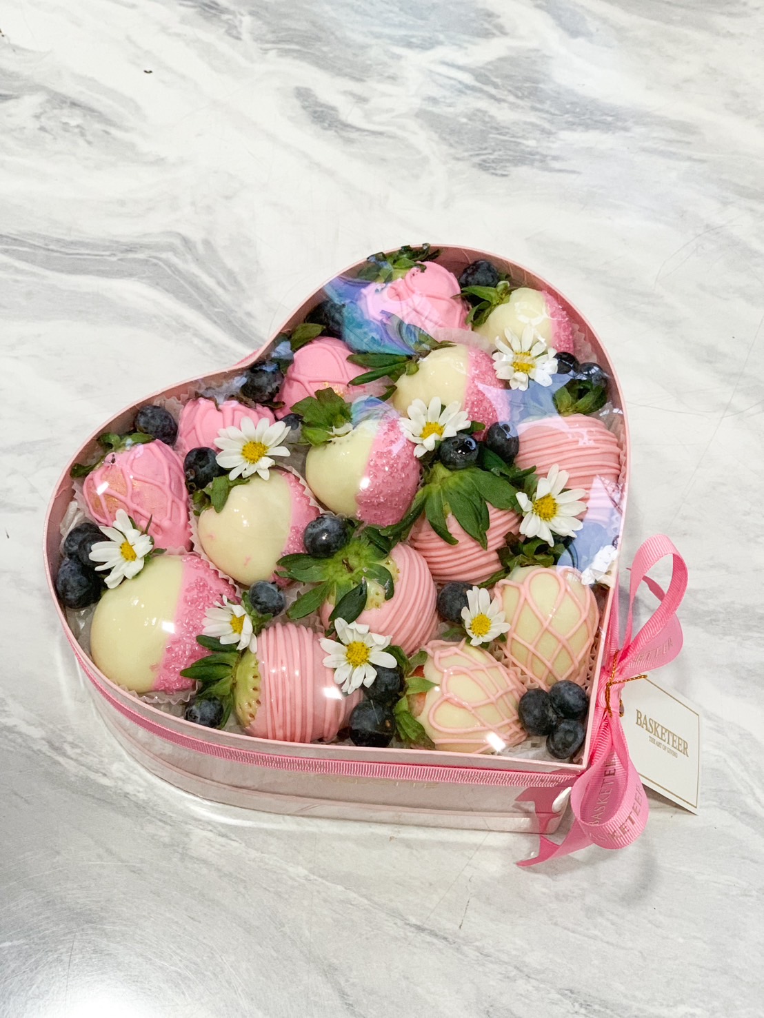 A heart-shaped pink box filled with pink and white chocolate-covered strawberries, blueberries, and small white flowers, creating an elegant and romantic dessert gift.