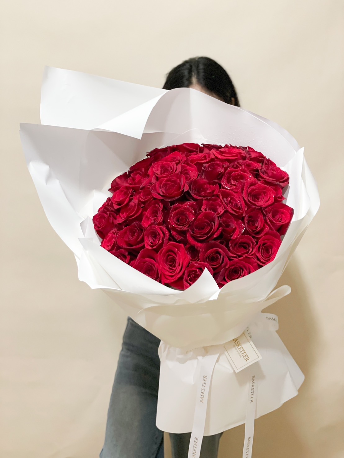 A luxurious oversized bouquet of 100 fresh red roses, elegantly wrapped in white with Basketeer branding, making it a perfect romantic gift for Valentine’s Day or anniversaries.