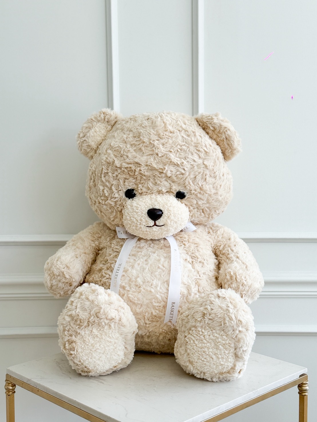 A large, fluffy beige Giant Plush Teddy Bear sits with a white ribbon round its neck on a white tabletop in front of a white panelled wall, perfect for adding the Giant Plush Teddy Bear Set to your collection.