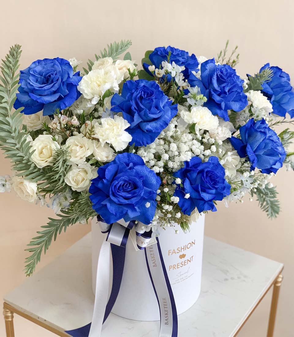 Close-up of a stunning floral arrangement featuring vibrant blue roses, delicate white flowers, and lush greenery, elegantly presented in a luxury gift box.