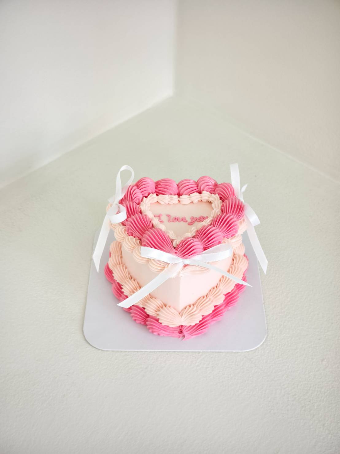 A heart-shaped Charlotte cake with pink and white icing, piped borders, white ribbons, and ‘I love you’ in pink icing on top.