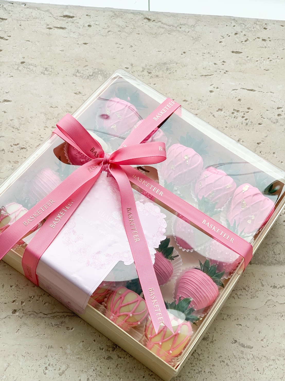 Elegant gift box of strawberries dipped in pink and white chocolate with decorative drizzle and gold accents, tied with pink ribbon.