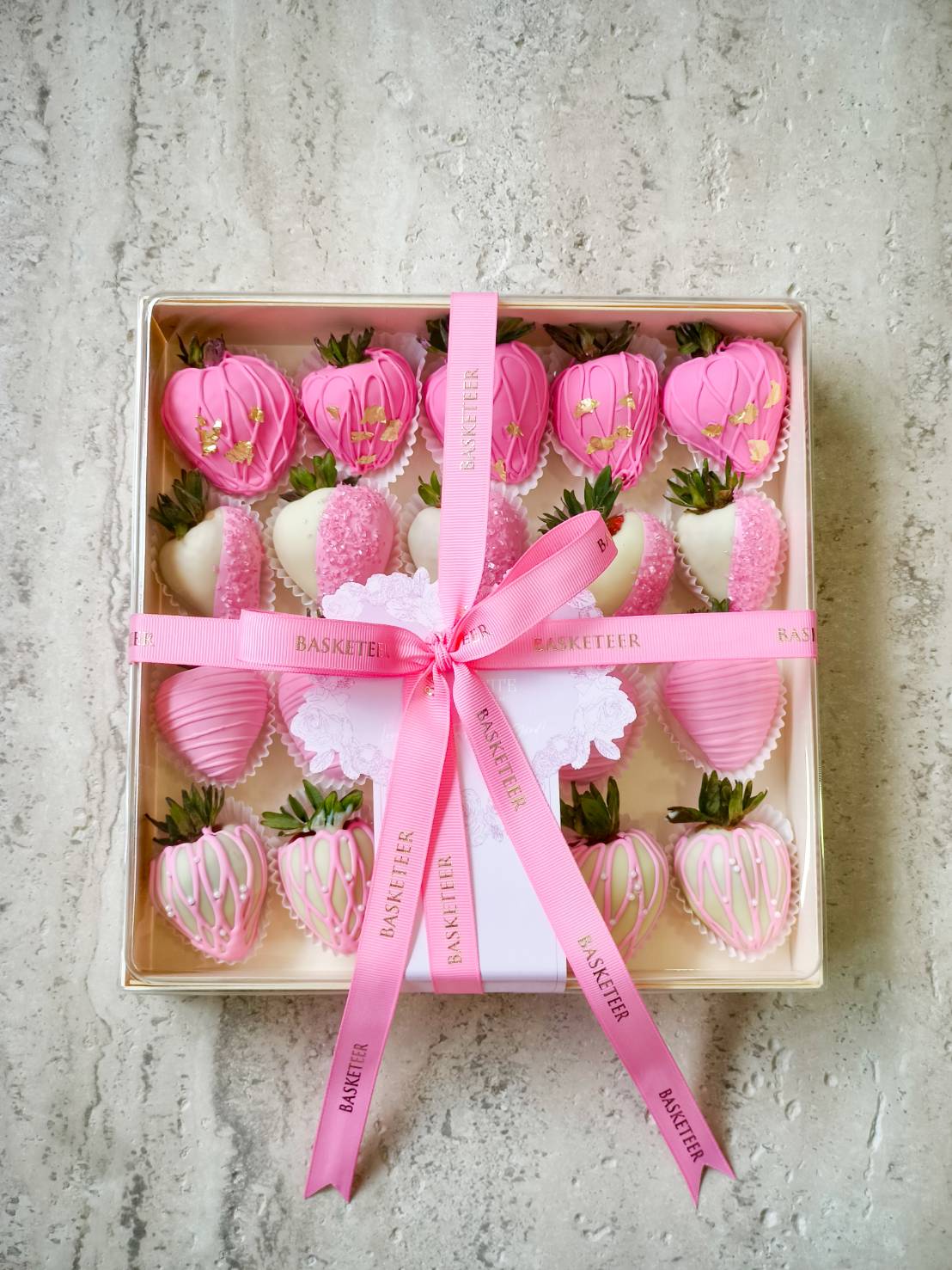 A luxury gift box of pink and white chocolate-covered strawberries decorated with gold leaf, sprinkles, and elegant drizzle designs.