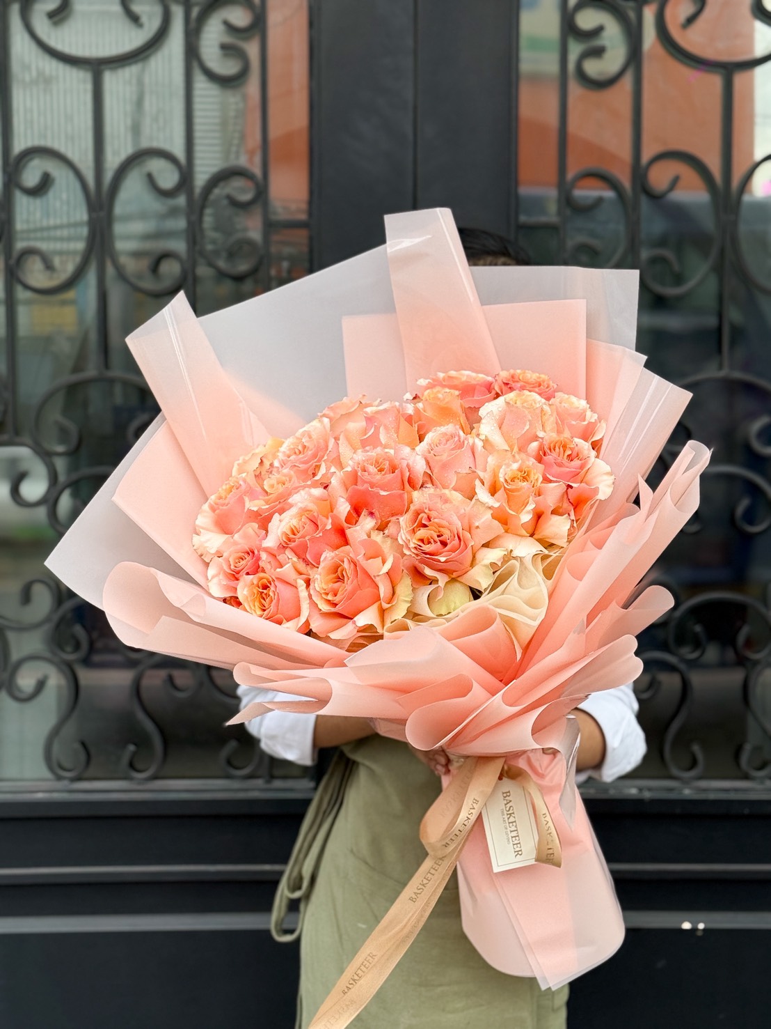 A person in an apron holds a stunning Peach Rose Bouquet of peach and pink roses, wrapped in peach and beige paper, standing before an ornate black iron door.