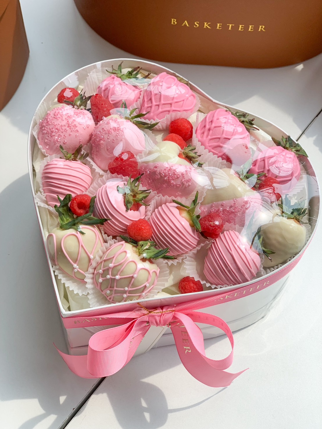 A heart-shaped gift box filled with pink and white chocolate-covered strawberries, decorated with drizzles and raspberries.