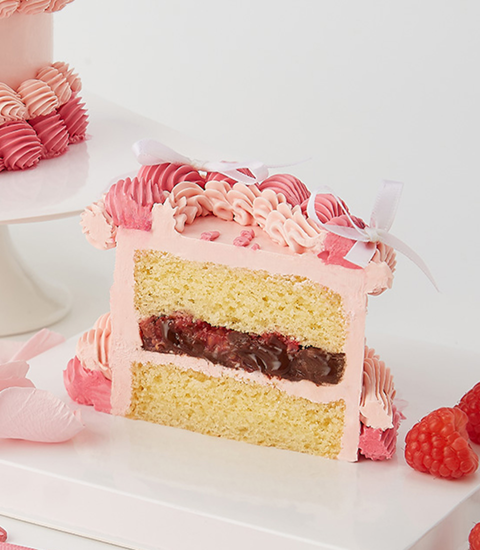 A slice of pink frosted cake with vanilla sponge layers, raspberry chocolate filling, and intricate buttercream piping.