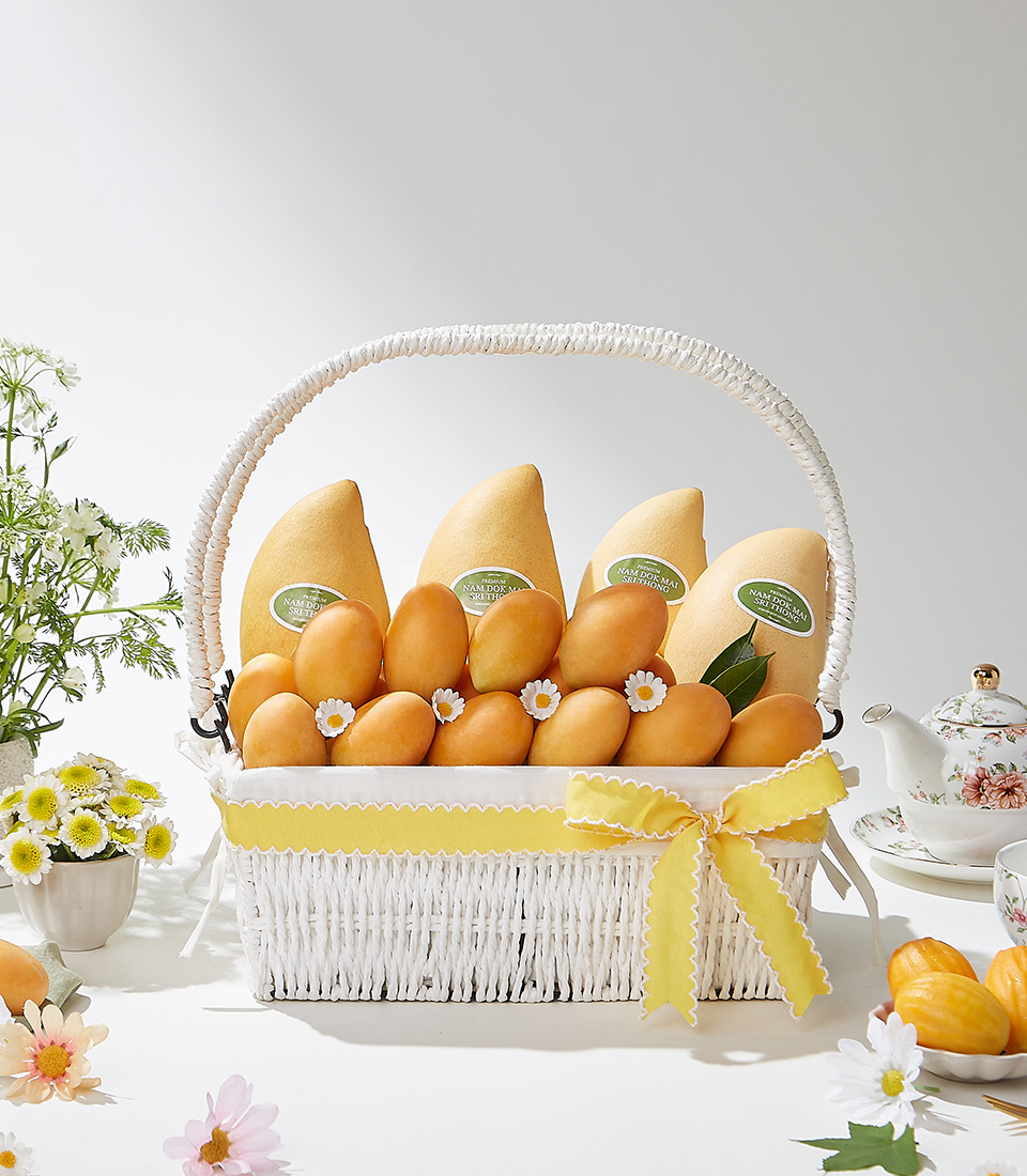 A sophisticated white wicker basket filled with premium Toon Klaow variety Mayongchid and Nam Dok Mai Barracuda Mangoes, elegantly adorned with a yellow satin ribbon and delicate floral accents.