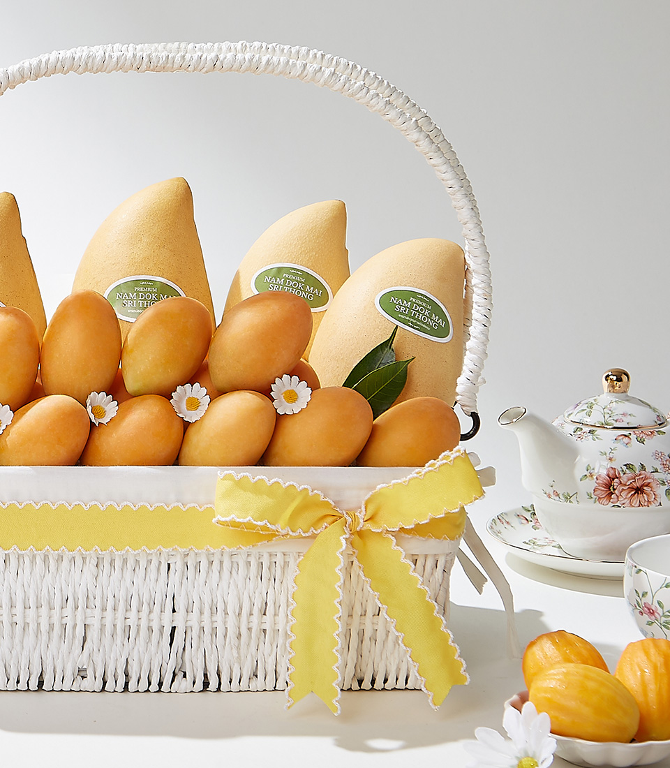 A sophisticated white wicker basket filled with premium Toon Klaow variety Mayongchid and Nam Dok Mai Barracuda Mangoes, elegantly adorned with a yellow satin ribbon and delicate floral accents.