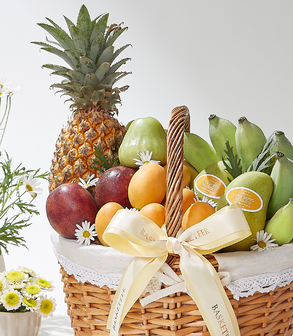 A wicker basket filled with premium Mayongchids from Nakhon Nayok, Ok-Rong mangoes from Damnoen Saduak, Pattawia pineapple, passion fruits, green Cavendish bananas, and green rose apples elegantly decorated with a cream ribbon and white lace