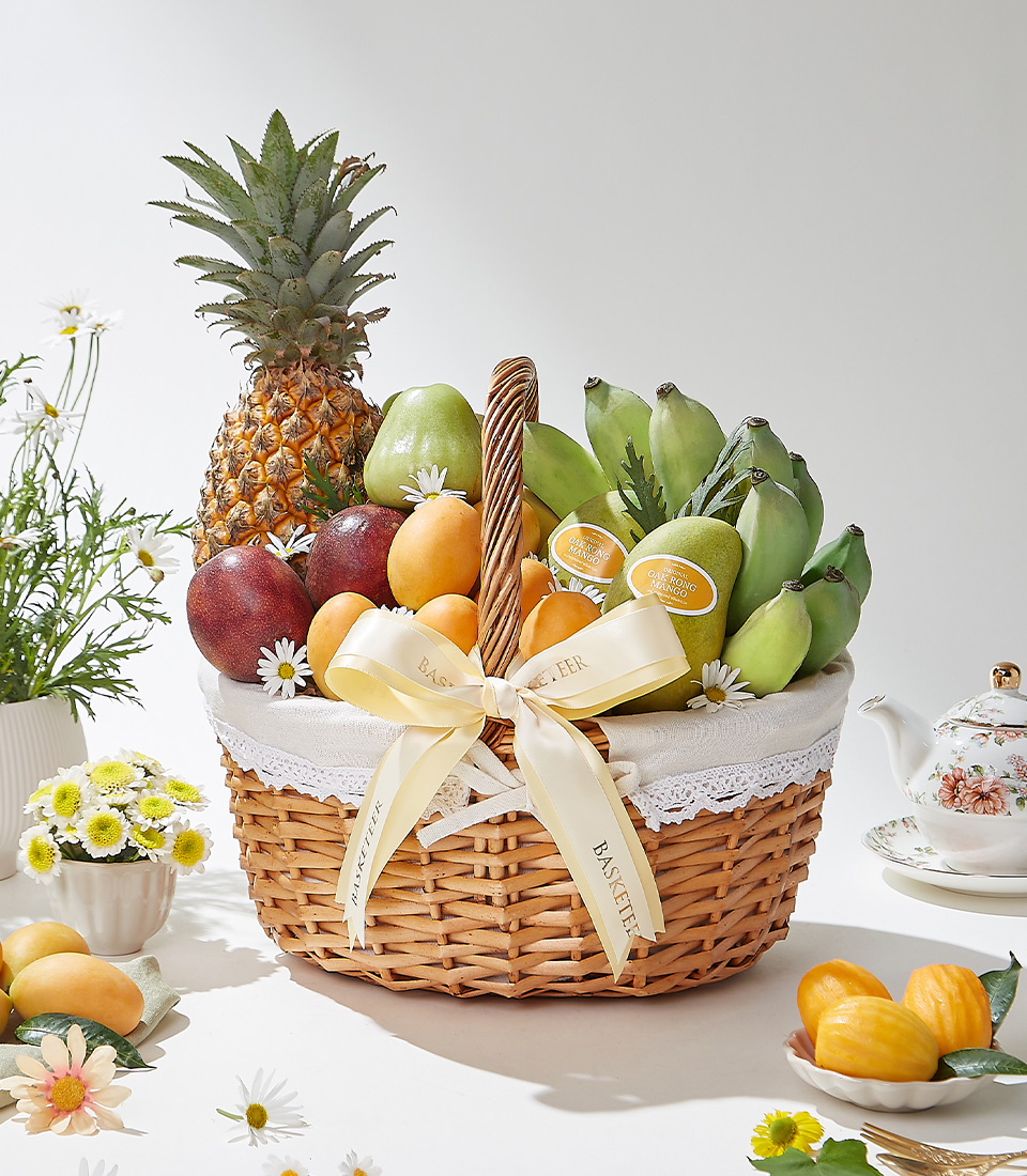 A wicker basket filled with premium Mayongchids from Nakhon Nayok, Ok-Rong mangoes from Damnoen Saduak, Pattawia pineapple, passion fruits, green Cavendish bananas, and green rose apples elegantly decorated with a cream ribbon and white lace