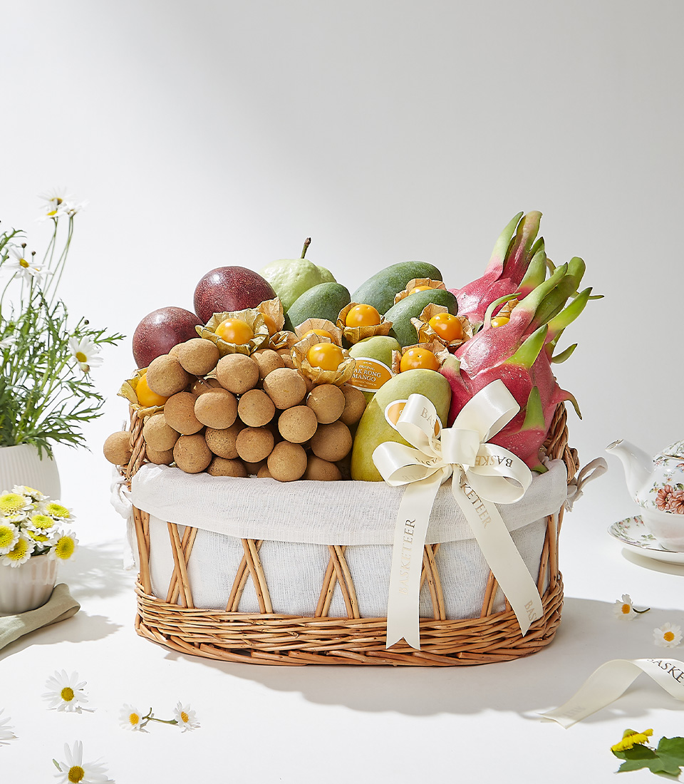 A wicker basket filled with Ok-Rong mangoes from Damnoen Saduak, Keo Seowei green mangoes, Kimju guava, red skin dragon fruits, passion fruits, longans, and cape gooseberries beautifully arranged with a cream ribbon and white cloth lining